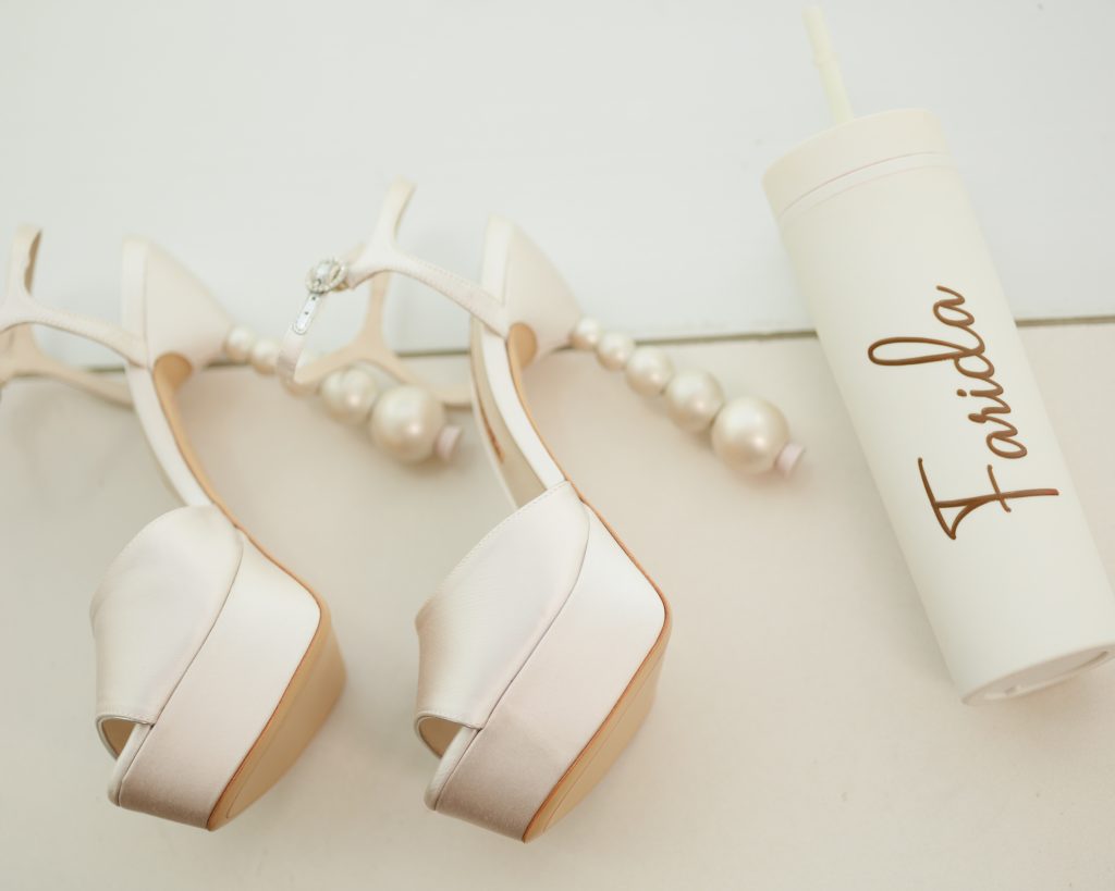 White platform heels with pearl accents beside a personalized bridal cup reading “Farida.”